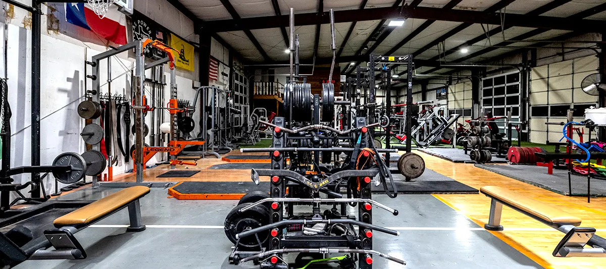 A wide view of the gym floor: turf, machines, benches and platforms under a high metal roof with daylight through the roll-up doors.